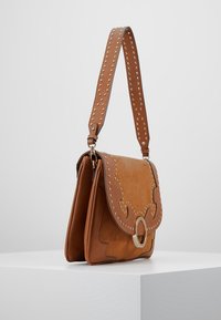 Hnědá kožená taška přes rameno s akcenty zlatých hřebíčků, dekorativním vlnitým designem a kruhovým zapínáním. Nastavitelný popruh se hřebíčky.