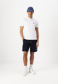White polo shirt with small logo, navy drawstring shorts with side pocket, white sneakers, and blue baseball cap. Simple, casual outfit.