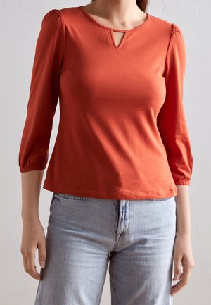 Rotes Baumwolloberteil mit 3/4 Puffärmeln, Ausschnitt mit Schlüsselchen-Detail und taillierter Silhouette. Kombiniert mit hochgeschnittenen hellblauen Jeans.
