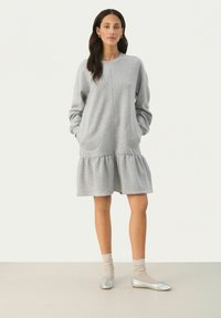 Graues Sweatkleid mit langen Ärmeln, seitlichen Taschen und einem gestuften Rüschensaum. Trägt man mit silberfarbenen Ballerinas und beigen Socken. Glatte Textur.