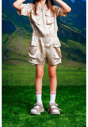 Combinaison courte en satin beige avec manches courtes, fermeture éclair et deux poches avant. Associée à des chaussettes blanches et des sandales beiges avec des accents rouges.