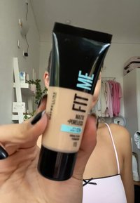 Beige tub av Maybelline Fit Me Matte + Poreless foundation med svart kork, med blå och svart text, hålls i en hand.
