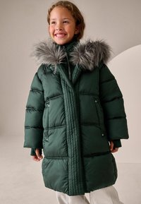 Zelena puffer jakna s umjetnim krznom na kapuljači, u kvadratnom uzorku, i zatvaranjem na patentni zatvarač. Ima široke rukave i prednje kopčanje na dugmadi.
