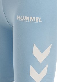 Jasnoniebieska tkanina sportowa z białym napisem "HUMMEL" oraz dwoma białymi logo w kształcie szewronów skierowanych w dół na obszarze uda.