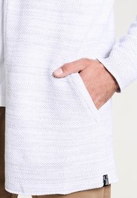 Hvid strikket cardigan med en tekstureret overflade, der har en sidelomme og et mærke på bundkanten. Glat stof, afslappet pasform.