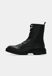 Crna kožna borbena čizma s dizajnom na vezanje, visokim gležnjem i debelim gumenim potplatom. Ima metalne rupice za vezice i trakicu za povlačenje na stražnjoj strani.