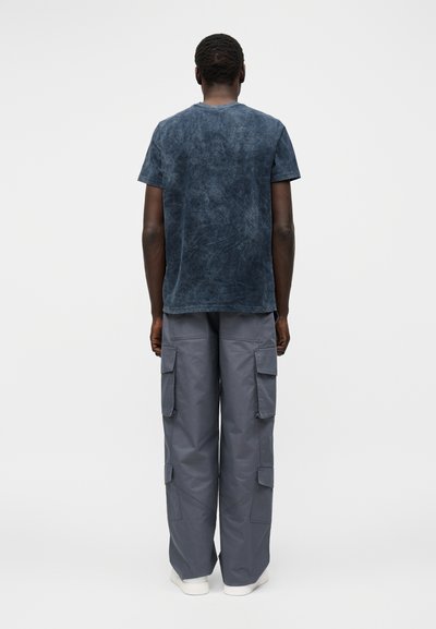 Άνδρας με την πλάτη φοράει μια κοντομάνικη, σκουρόχρωμη μπλε φανέλα πλυμένη και φαρδιά γκρι παντελόνια cargo με μεγάλες πλαϊνές τσέπες και λευκά παπούτσια.