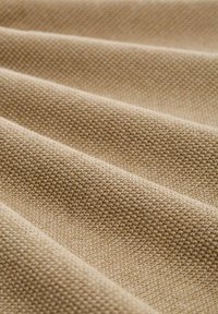 Beige strukturiertes Gewebe mit einem gewebten Muster, das weiche Falten und eine glatte Oberfläche aufweist. Das Material wirkt dick, aber dennoch flexibel.