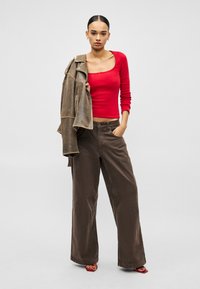 Crvena majica dugih rukava s kvadratnim izrezom, uparena s širokim smeđim hlačama i smeđom kožnom jaknom, prikazujući ležerni kroj.