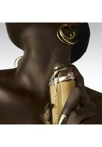 Flacon de parfum doré avec un bouchon en forme de diamant, tenu par une main aux ongles longs et verts. Peau lisse et boucles d'oreilles circulaires dorées visibles.