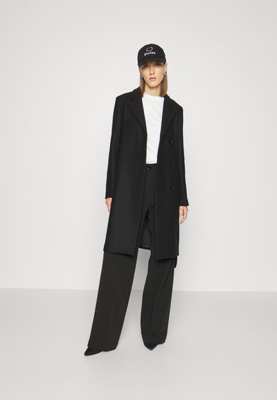 Black long coat in a smooth fabric, double-breasted design, worn over a white shirt and wide black trousers, complemented by a black cap.