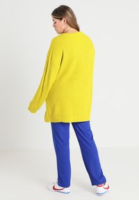 Gul ribbet oversized genser med lange ermer, kombinert med blå bukser. Hvite sko med røde aksenter fullfører antrekket.