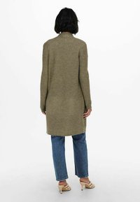 Cardigan lungo a coste in verde oliva, con colletto alto e apertura frontale. Abbinato a jeans azzurri dritti e scarpe con tacco beige.