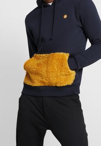 Marinblå hoodie med en stor, fluffy, gul ficka. Tyget är mjukt med en huva med dragsko och ribbade ärmslut, som kompletterar svarta byxor.