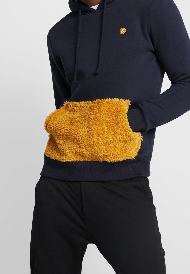 Marinblå hoodie med en stor, fluffy, gul ficka. Tyget är mjukt med en huva med dragsko och ribbade ärmslut, som kompletterar svarta byxor.