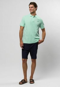 Világoszöld pólóing, rövid ujjú, galléros, bal oldalon logó, sötétkék rövidnadrággal és barna szandállal párosítva. Sima pamut felület.