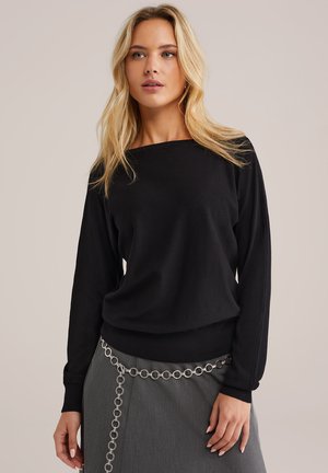Crni dugi pulover s čamastim izrezom, fino pletena tekstura, uparen s sivom suknjom koja ima srebrni lanac kao detalj.