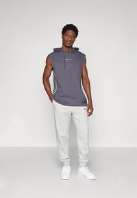 Man draagt een mouwloos donkergrijs hoodie en lichtgrijze joggingbroek met witte sneakers, staand met handen in de zakken.