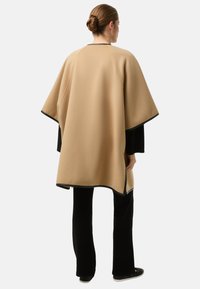 Beige poncho med glat tekstur og sort kant, der har en løs silhuet og afrundede kanter, båret over sorte, tætsiddende bukser.