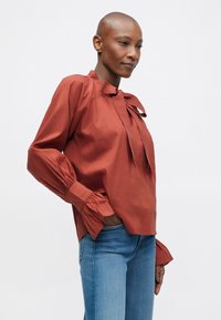 Camicia rossa a maniche lunghe con dettaglio a fiocco sul colletto, fronte plissettato e polsini arricciati. Indossata con jeans blu. Tessuto simile al cotone.