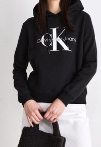 Černá mikina s kapucí s předním logem, s kapucí na šňůrku a žebrovanými manžetami, doplněná o texturovanou tmavě hnědou tašku.