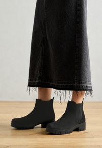 Svarta kängor i ankelhöjd gjorda av texturerat material med en klumpig sula och elastiska sidopaneler, kombinerade med en lång grå kjol med fransad fåll.