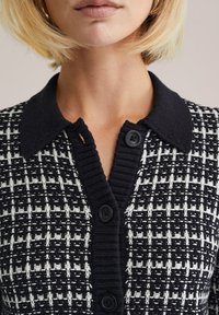 Pletený kardigán so vzorom čierno-bielych kociek, textúrovaným čiernym golierom a tromi čiernymi gombíkmi pozdĺž predného otvoru.