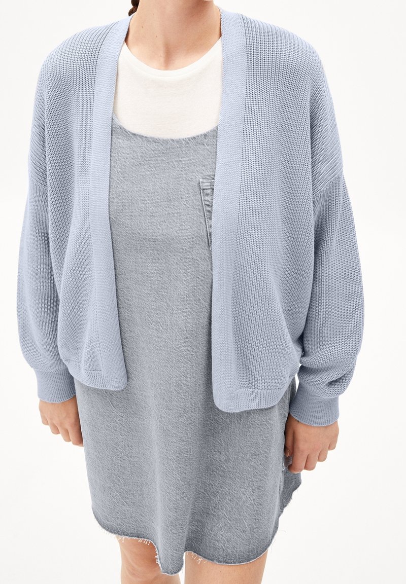 Vaaleansininen neulottu neuletakki, jossa on leveät hihat, käytetty harmaan hihattoman mekon päällä. Mekossa on teksturoitu pinta ja viimeistelemätön helma.