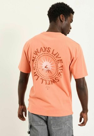 Mann trägt ein pfirsichfarbenes T-Shirt mit Sonnen- und Muschelmotiv sowie dem Text "Always live like a shell" auf dem Rücken, kombiniert mit grauen Shorts.