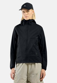 Schwarze wasserdichte Jacke mit durchgehendem Reißverschluss, Kapuze und elastischen Bündchen. Mit Logo auf der Brust. Das Modell kombiniert sie mit hellbeigen Hosen.