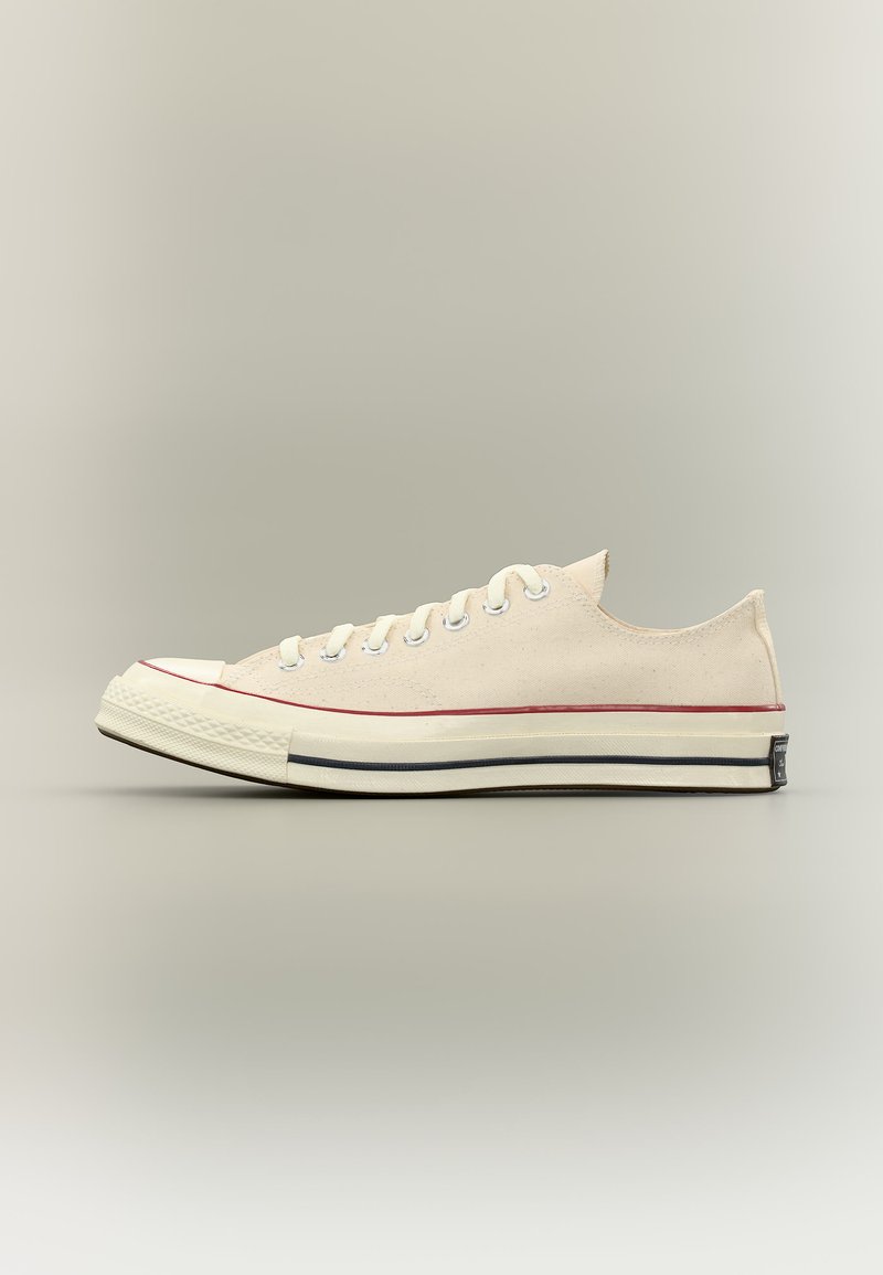 Sneaker beige in tela con taglio basso, lacci bianchi, punta in gomma e suola bianca con strisce rosse e nere.