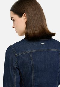 Džínová bunda v tmavě modré barvě s mírně strukturovaným střihem, viditelným šitím, černým límcem a kovovým logem na zadní straně.
