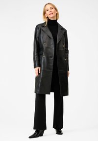 Vrouw die staat in een zwarte leren dubbelbreasted jas, zwarte turtleneck, zwarte broek en zwarte puntige laarzen op een witte achtergrond.
