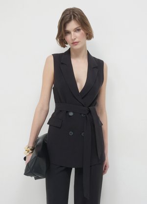 Blazer nero senza maniche con scollo a V profondo, doppiopetto con bottoni, cintura abbinata e tasche laterali, indossato sopra pantaloni neri.