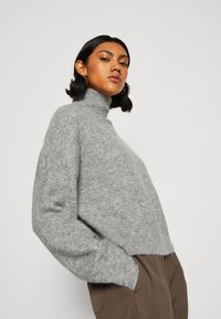 Pull en maille gris avec col roulé, coupe décontractée, texture douce et poignets côtelés. Associé à un pantalon marron, présenté sur fond blanc.