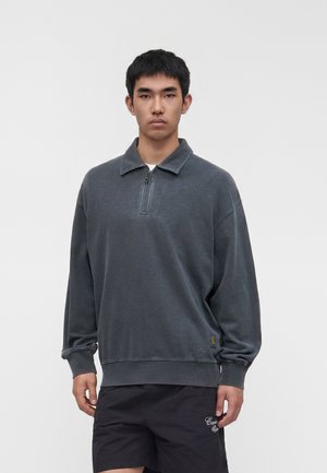 Ung mand iført en mørkegrå pullover med kvart lynlås og sorte shorts står foran en ensartet lys baggrund.