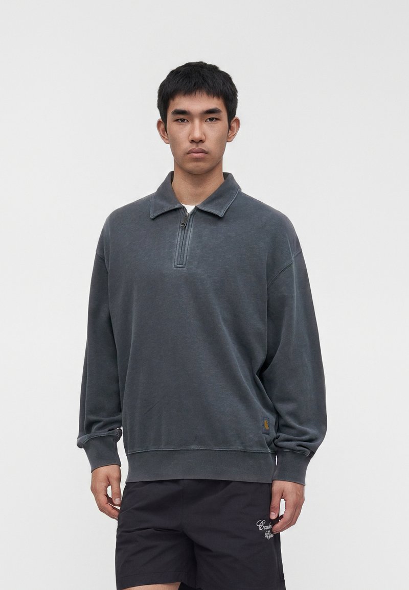 Young man wearing a dark gray quarter-zip pullover and black shorts standing against a plain light background.