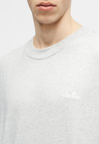 Κοντινό πλάνο ατόμου που φοράει ανοιχτό γκρι φούτερ ellesse με ριμπ λαιμόκοψη και μικρό λογότυπο της μάρκας στο στήθος.