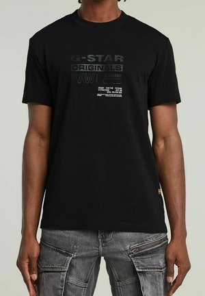 Homme portant un t-shirt graphique noir G-Star Originals et un jean cargo gris, debout devant un fond clair uni.