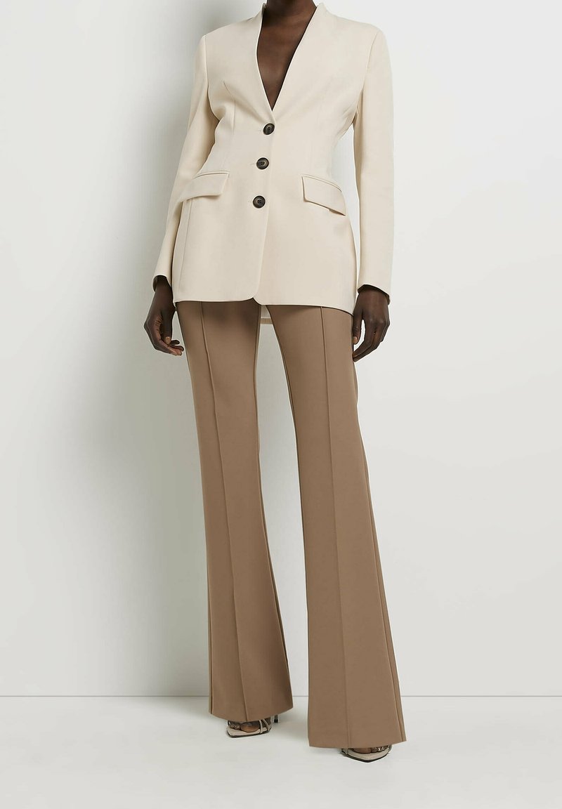 Personne à la peau foncée portant un blazer crème à trois boutons et un pantalon évasé taille haute marron clair, debout contre un mur blanc.