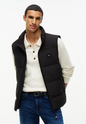 Ung mand iført en sort dunvest over en hvid sweater med krave og blå jeans, stående mod en ensfarvet hvid baggrund.