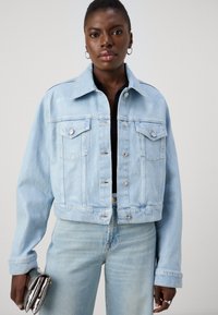 Svetlo modra denim jakna s skrajšanim dizajnom, dvema prsoma žepoma in srebrnimi gumbnimi detajli. Ustvarjena v kombinaciji s svetlo modrimi kavbojkami in kovinsko torbico.