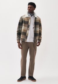 Jachetă cu model tartan în nuanțe de bej, maro și negru, dotată cu un guler din blană sintetică albă. Asortată cu o cămașă cu dungi deschise, pantaloni bej și adidași negri.