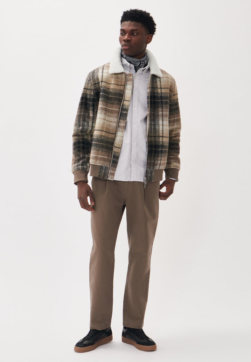 Jacke mit Karomuster in Beige, Braun und Schwarz, mit einem weißen Kunstfellkragen. Kombiniert mit einem hellgestreiften Hemd, beigen Hosen und schwarzen Sneakers.