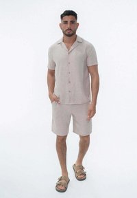 Chemise beige à manches courtes à boutons pour homme et short assorti, en tissu texturé, associés à des sandales marron à deux brides.