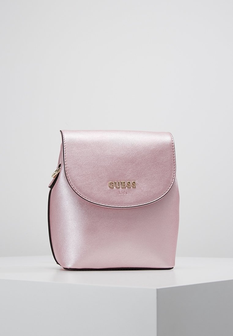 Vaaleanpunainen metallinhohtoinen reppu, valmistettu synteettisestä materiaalista. Siinä on pyöristetty läppä, kultainen logo ja tukeva musta pohja. Kompakti muotoilu ja tyylikäs pinta.