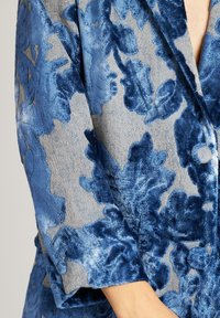 Tejido de terciopelo floral azul con una superficie texturizada, que presenta un suave brillo. El diseño incluye grandes y elaborados patrones de flores y acentos de botones.