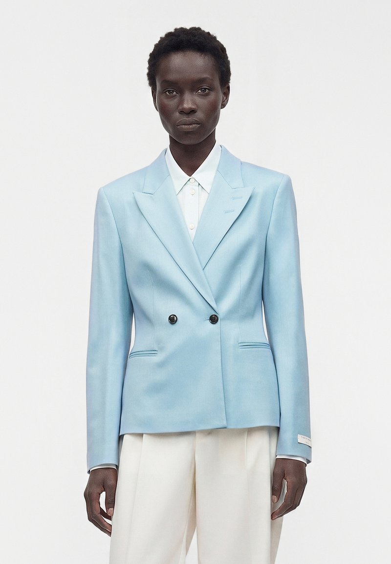 Dark-skinned person wearing a light blue double-breasted blazer over a white button-up shirt and white pants, against a plain background.