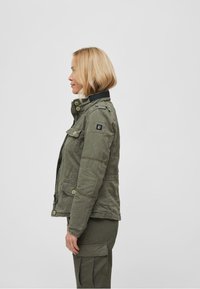 Olive green jacket with a zip and button closure, multiple pockets, and a black collar. Features a logo patch on the left sleeve.