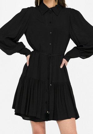 Mujer vestida con un vestido negro de manga larga con botones, cuello, cinturón ajustado en la cintura y falda plisada, las manos apoyadas en las caderas.